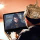 A man with a tinfoil hat sits alone in a room staring at his laptop which has an image of Bayonetta on his screen winking back at him.